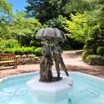 Bronze sculpture of two figures under an umbrella in a bright outdoor pool setting.