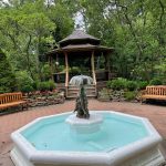 Stone fountain in a park setting with gazebo and lush green trees surrounding it.