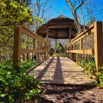 Wooden bridge leading to a gazebo in a sunlit, wooded park setting.