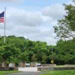 American flag flies over a landscaped park area with lush green trees under a cloudy sky.
