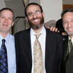 Three men in suits smiling together at an indoor event.