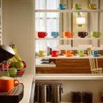 Kitchen counter with coffee maker and fruit, next to white shelving displaying colorful mugs.