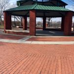 Green gazebo with brick base on a red brick plaza under a blue sky.