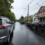 Gray SUV parked on a wet, tree-lined residential street under an overcast sky.