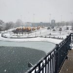 Snowy winter scene over a frozen pond with distant buildings under overcast skies.