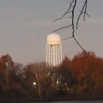 Water tower silhouetted against a pale sky, framed by bare tree branches and autumn foliage.