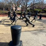 Bronze statues of children playing on a paved plaza with trees and buildings in the background.