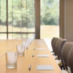 Long wooden conference table set up in a bright room with large windows overlooking greenery.