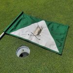 Green flag with a white and green design, next to a golf ball marker on artificial turf.
