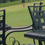 Golfer swings club on a green course with trees in the background.