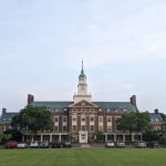 Historic brick university building with a central clock tower, set on a green lawn under a light sky.