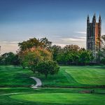 Gothic cathedral towers rise above a lush green golf course under a blue sky.