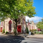 Historic stone church with red doors under a bright, sunny sky and lush green trees.
