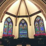 Stained glass windows illuminate a vaulted church interior with colorful religious scenes.