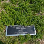 Sign at the base of lush green shrubbery: "The Awakening Seward Johnson."