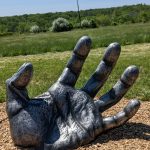 Giant gray hand sculpture resting on wood chips in a grassy, sunny outdoor field.