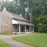 Stone farmhouse with white porch nestled among lush green trees.