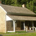 Stone cottage with a porch nestled among lush green trees on a sunny day.