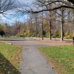 Autumn park scene with a pathway leading through trees and a stone wall on the left.