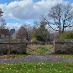 Stone wall with iron gate bordering a grassy area under a partly cloudy sky.