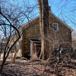 Old, weathered farmhouse under bare winter trees against a bright blue sky.