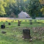 Cemetery with numerous old gravestones under a bright, leafy sky near a stone building.