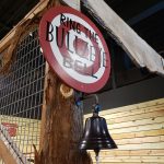 Red "RING THE BULLSEYE BELL" sign mounted on a wooden post with a hanging bell.