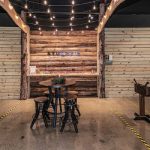 Rustic bar setup with a wooden backdrop, string lights, and small tables in an industrial space.