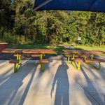 Picnic tables under a blue canopy in a sunny, wooded park setting.