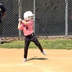 Baseball player in pink jersey swings bat during practice against a chain-link fence.