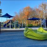 Bright playground with blue slides, green wavy climber, and autumn trees under a clear sky.