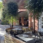 Outdoor dining area beneath a wooden overhang with greenery and brick architecture.