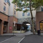 Outdoor dining area under an archway between modern brick and stone buildings.