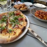 Outdoor table with pizza, pasta dish, bread, and drinks.