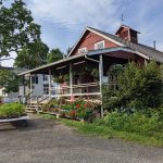 Red farmhouse with porch and garden beds under a blue sky.