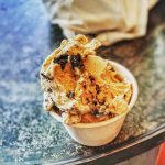 Scoop of coffee-flavored ice cream in a small paper cup on a textured surface.