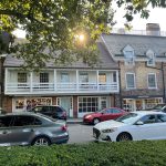 Historic brick buildings with storefronts under a bright, sunlit tree canopy.