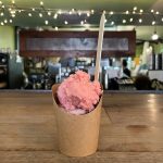 Pink scoop of ice cream in a brown paper cup on a wooden counter.