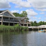 Large waterfront house with deck overlooks calm river under a partly cloudy sky.