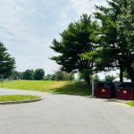 Paved driveway leading past outdoor trash and recycling bins near green trees.
