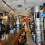 Brewery interior with large stainless steel fermentation tanks and wooden bar counter.