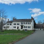 Historic white farmhouse under a blue, cloudy sky with a paved driveway leading away.