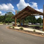 Wooden pavilion shelter over picnic tables with trees and blue sky background.