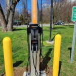 Electric vehicle charging station with yellow bollards in a park setting under a blue sky.