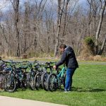 Man in black jacket stands near a row of parked bicycles on a grassy park path.