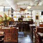 Rustic, modern restaurant interior with warm lighting, wooden tables, and large floral centerpiece.