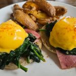 Breakfast plate with avocado toast topped with sunny-side-up eggs and spinach, served with fried plantains.