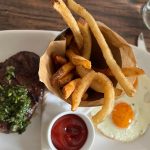 Steak, french fries in a cone, and fried egg on a white plate.