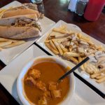 Sandwiches, french fries with drizzle, and a bowl of soup on a dark wooden table.