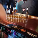 Bar counter with beer glasses and bottles, people socializing in a dimly lit pub setting.